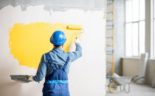 Workman in uniform painting wall with yellow paint at the construction site indoors