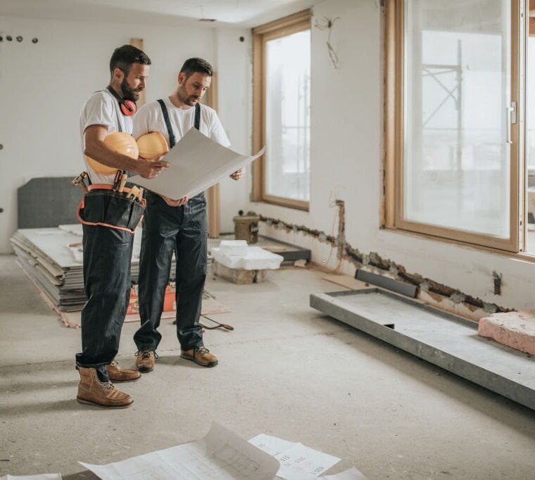 Young manual workers standing at construction site and examining blueprints.