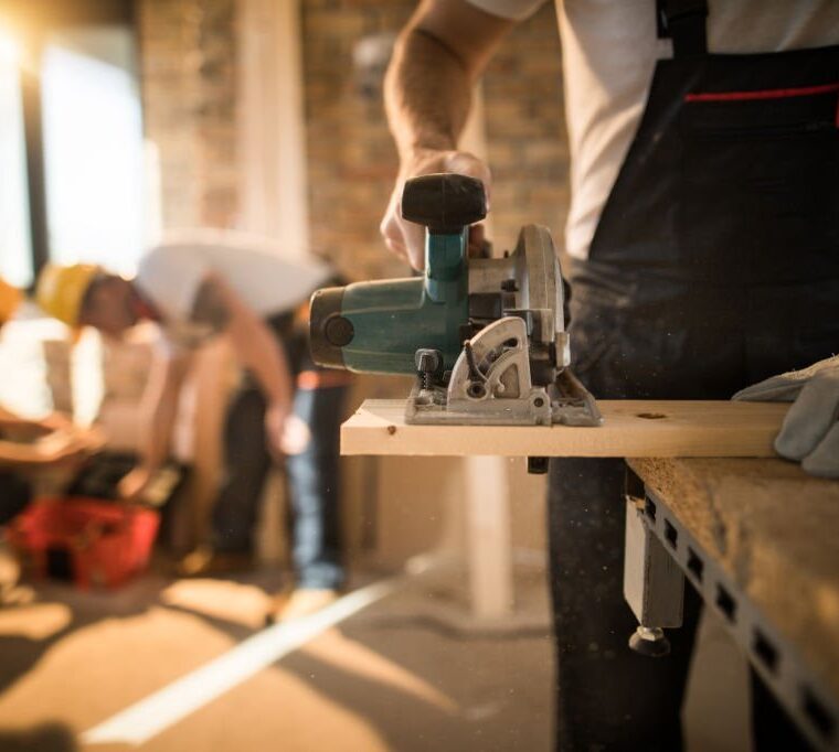 Unrecognizable manual worker using circular saw while cutting piece of wood at construction site. His colleagues are in the background.